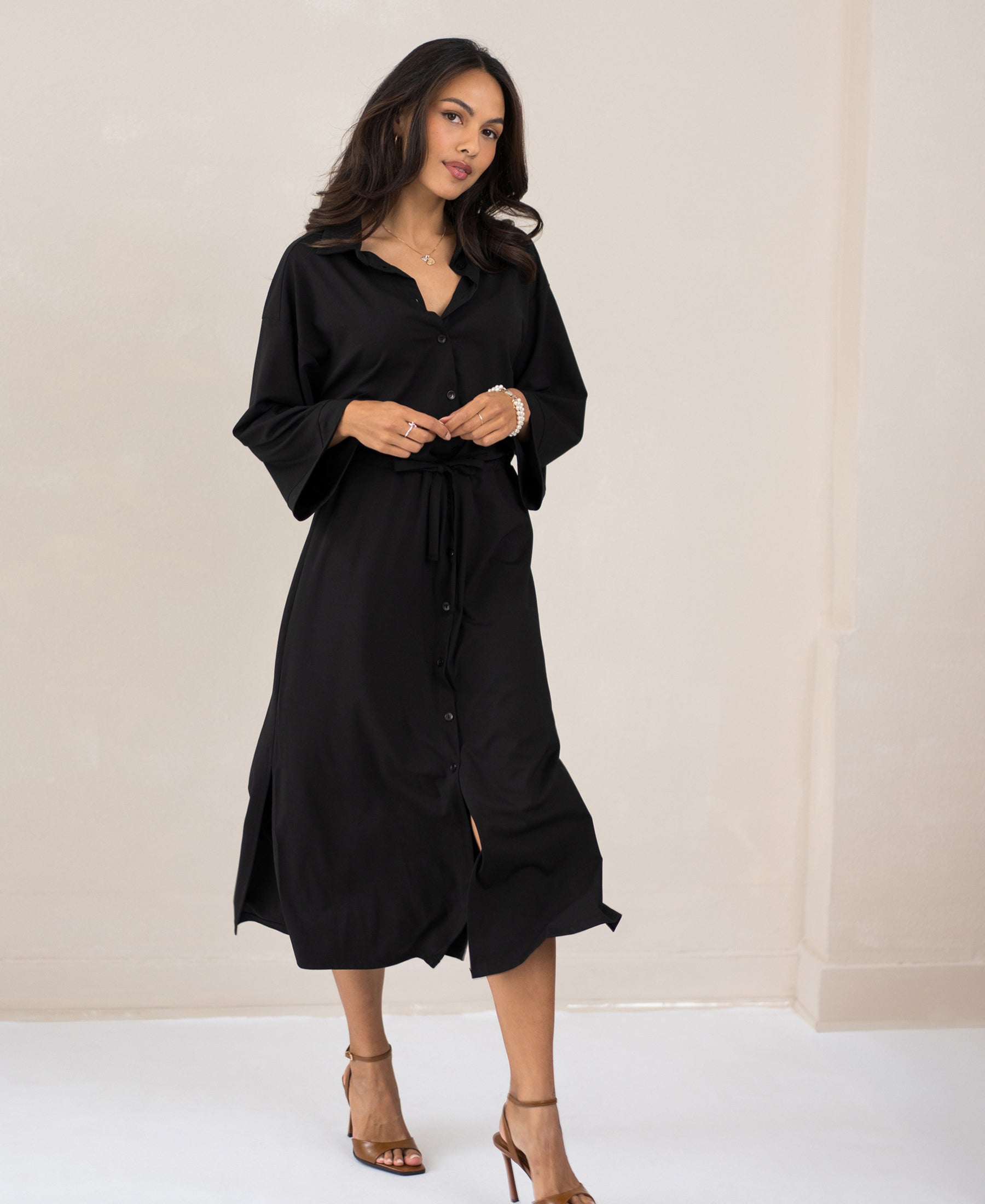 Woman wearing black LE RAPHAEL button-down dress with kimono sleeves by PLEIN PUBLIQUE, standing indoors.