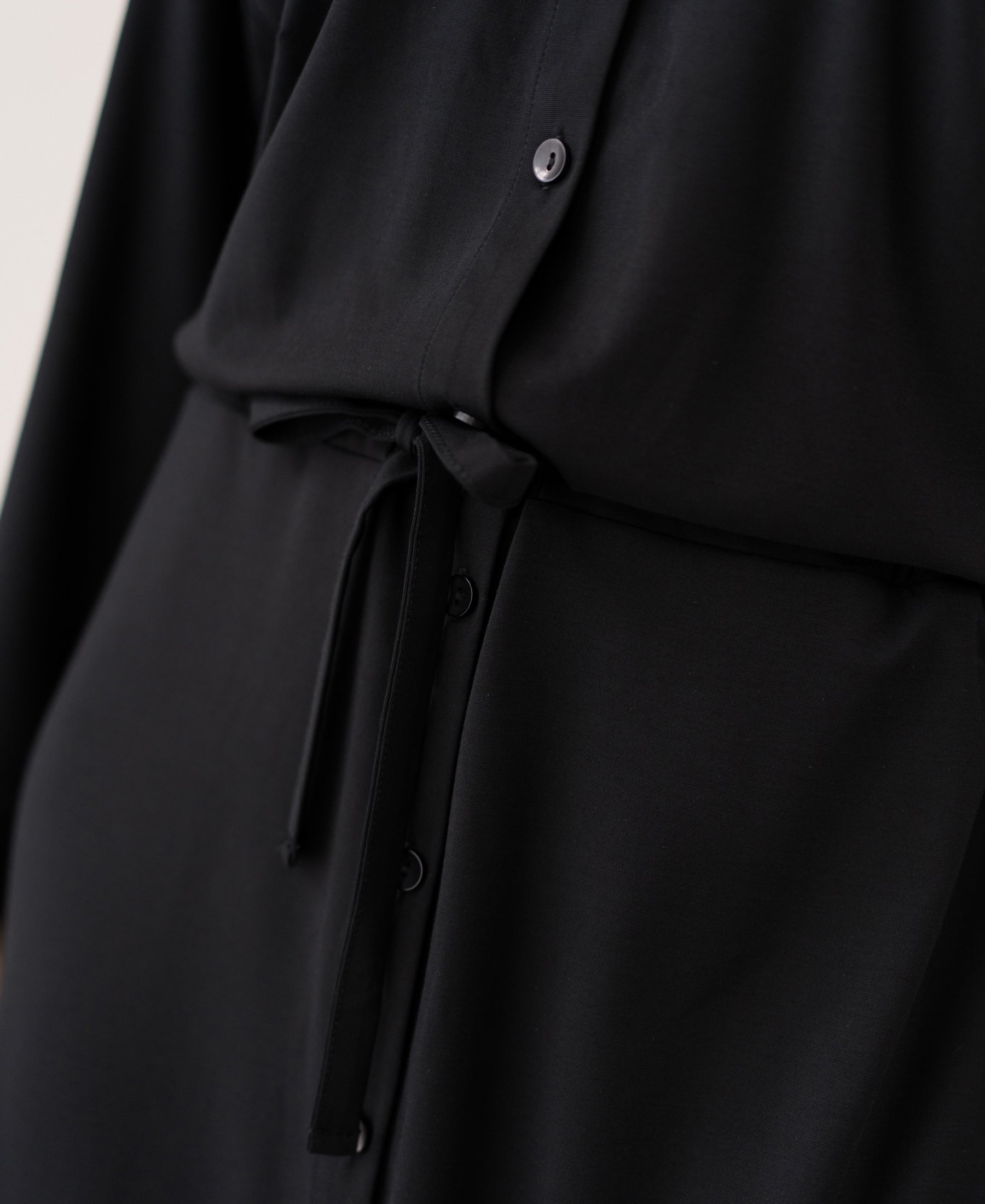 Close-up of the LE RAPHAEL Black Button-Front Dress by PLEIN PUBLIQUE, showing smooth fabric and a waist tie.
