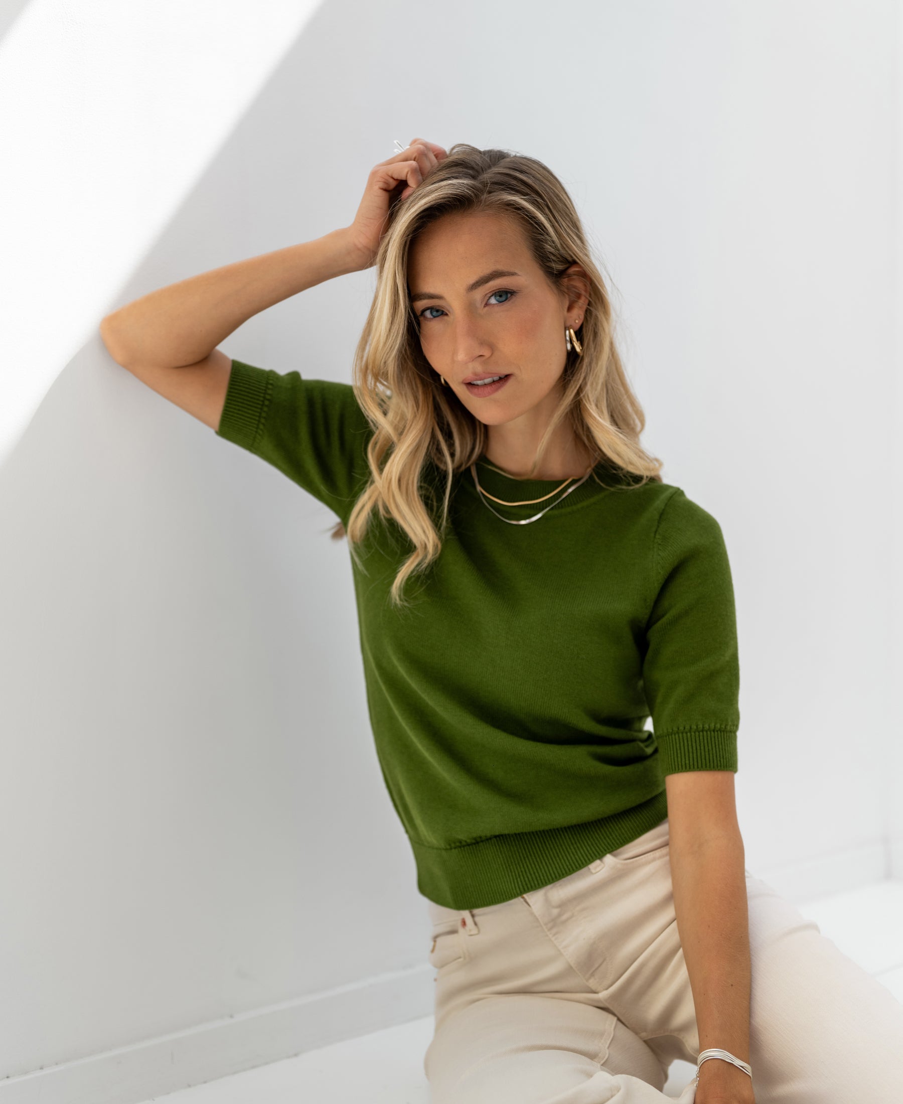 Woman wearing an olive green LA REVE sweater by PLEIN PUBLIQUE, sitting in natural light.