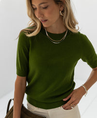 Woman wearing an olive green "LA REVE" sweater from PLEIN PUBLIQUE, looking down against a white background.