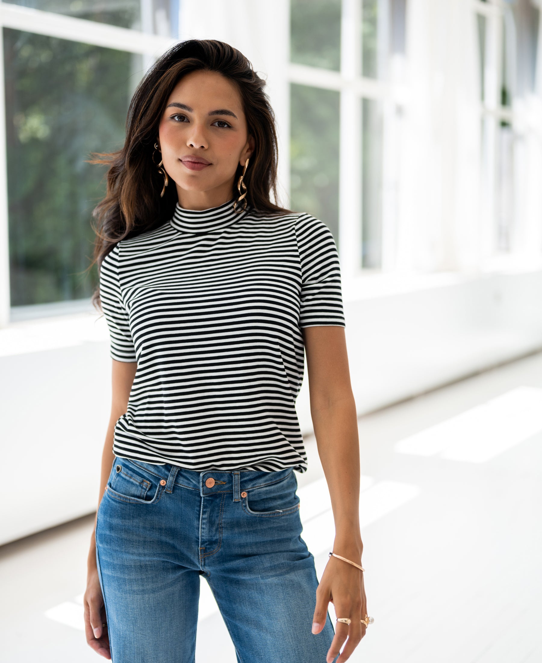 A woman wears a white and black ribbed collared top by PLEIN PUBLIQUE in a sunlit room with large windows.