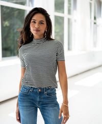A woman wears a white and black ribbed collared top by PLEIN PUBLIQUE in a sunlit room with large windows.
