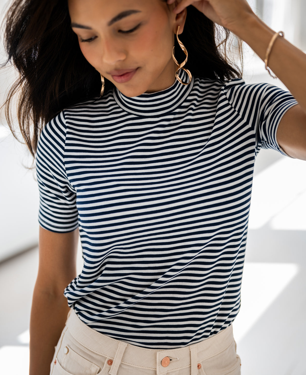 Woman wears the PLEIN PUBLIQUE ribbed collared top in white-dark blue, smiling softly and holding an earring.