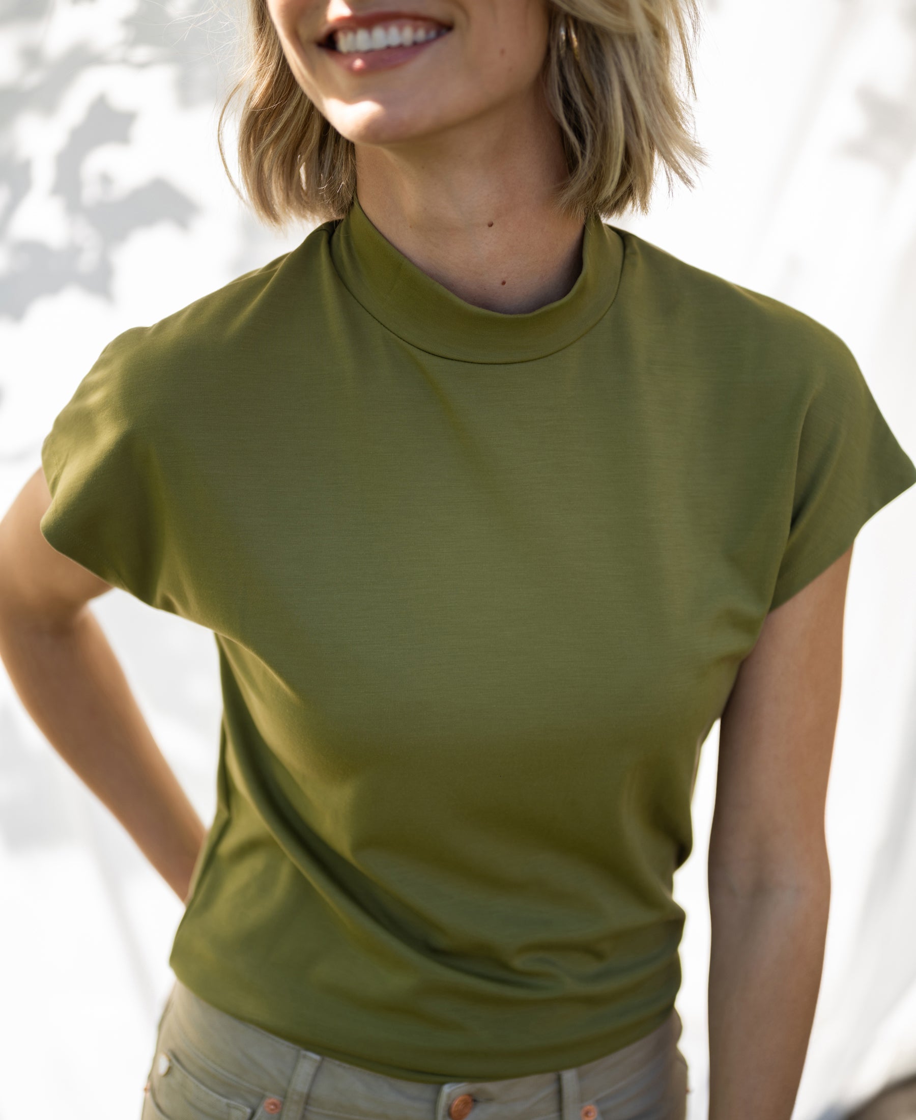 Olive green LA ROMY travel blouse by PLEIN PUBLIQUE shown in sunlight, with a softly blurred white background.