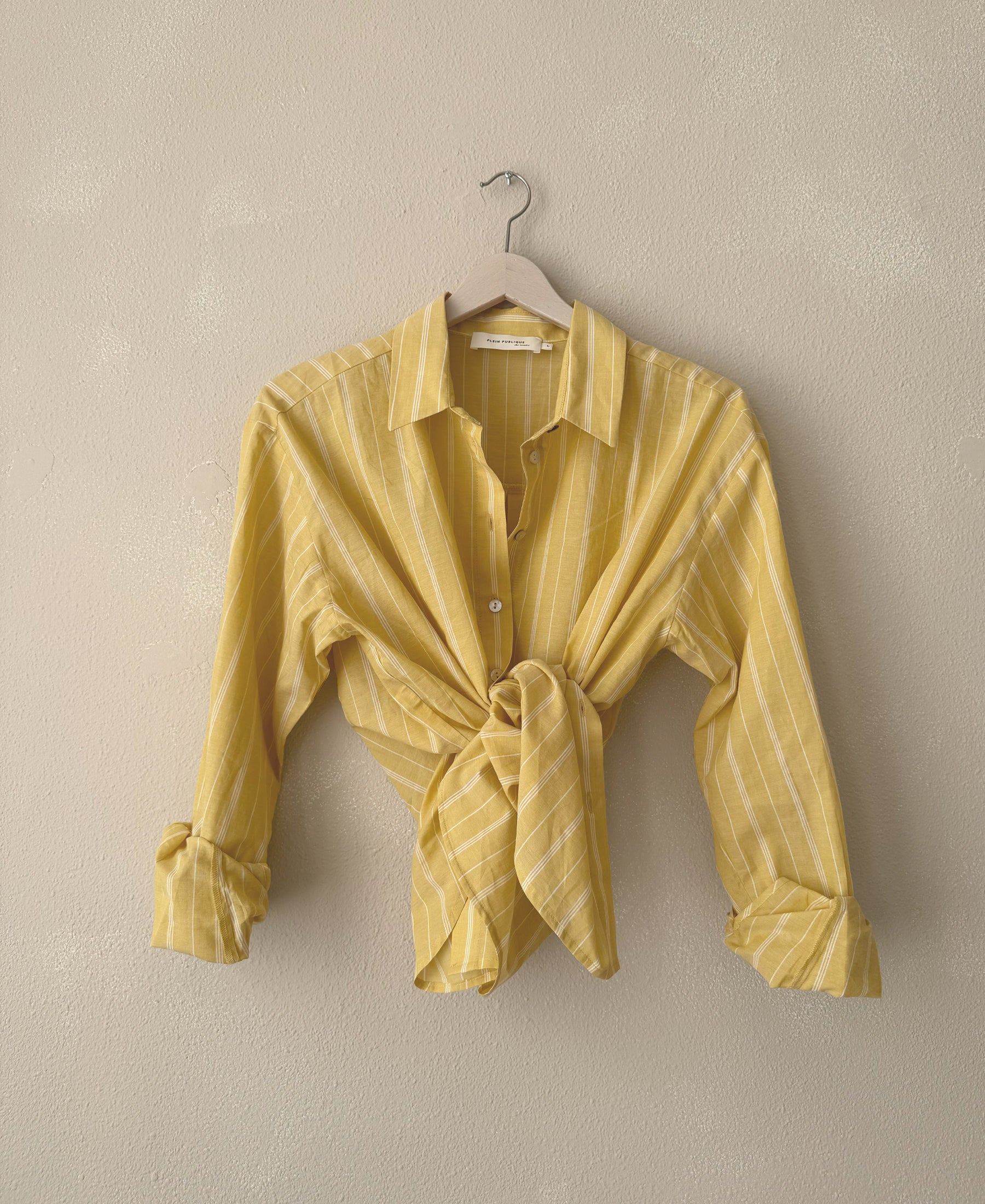 Oversized yellow striped blouse, tied at waist, hanging against a beige wall.
