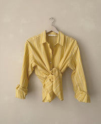 Oversized yellow striped blouse, tied at waist, hanging against a beige wall.