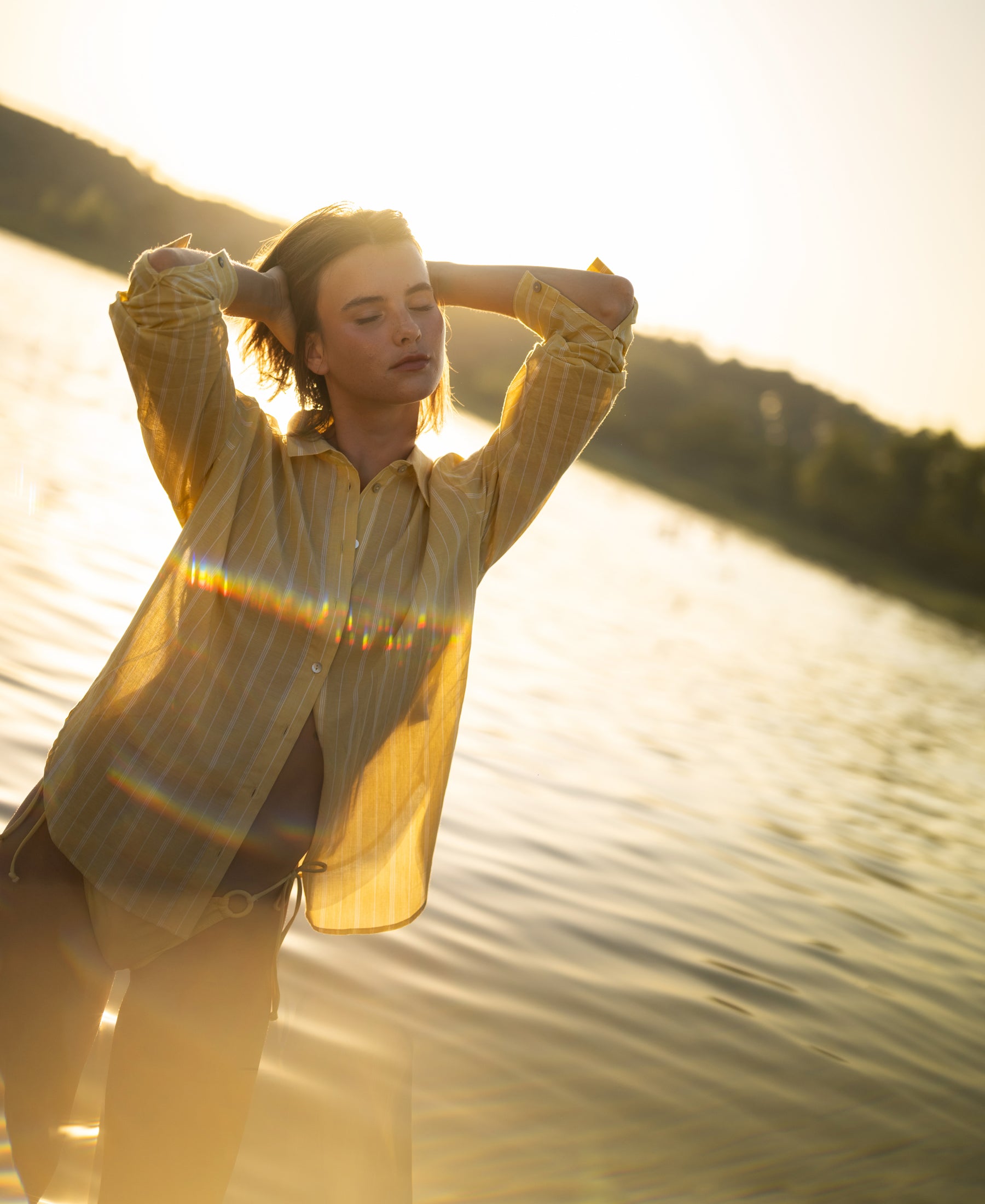 Oversized yellow striped blouse by PLEIN PUBLIQUE, front view, worn outdoors at sunset with arms raised.