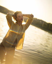 Oversized yellow striped blouse by PLEIN PUBLIQUE, front view, worn outdoors at sunset with arms raised.