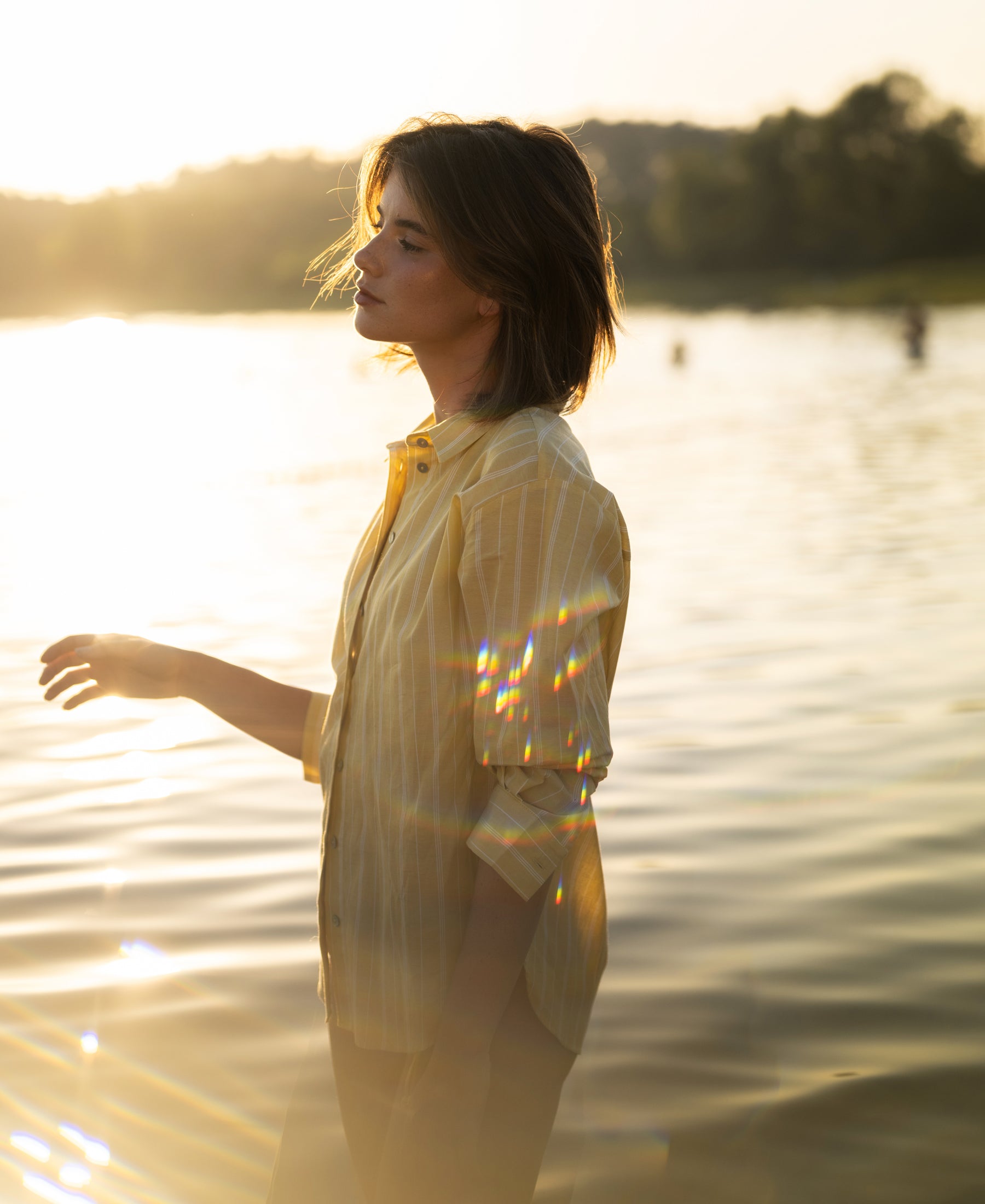 Oversized yellow striped blouse by PLEIN PUBLIQUE, shown in shallow water at sunset with sunlight glimmering.