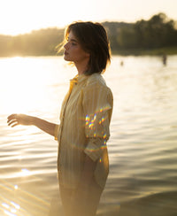 Oversized yellow striped blouse by PLEIN PUBLIQUE, shown in shallow water at sunset with sunlight glimmering.
