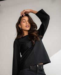 Woman with long wavy hair wearing a PLEIN PUBLIQUE pinstripe LE SAINT top in black, eyes closed, hand raised.