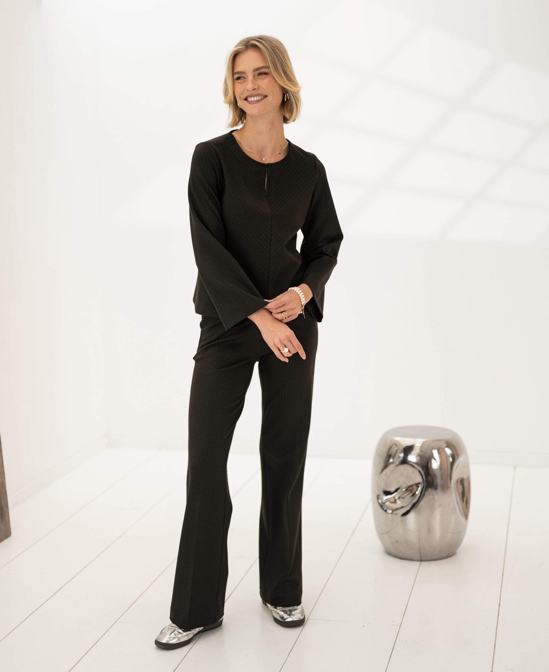 Woman wearing striped top in pure chocolate color, standing in bright white room near metallic stool.