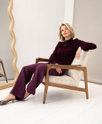 Wide-leg trousers in wine color, shown worn by model seated in a beige armchair; wavy mirror in the background.