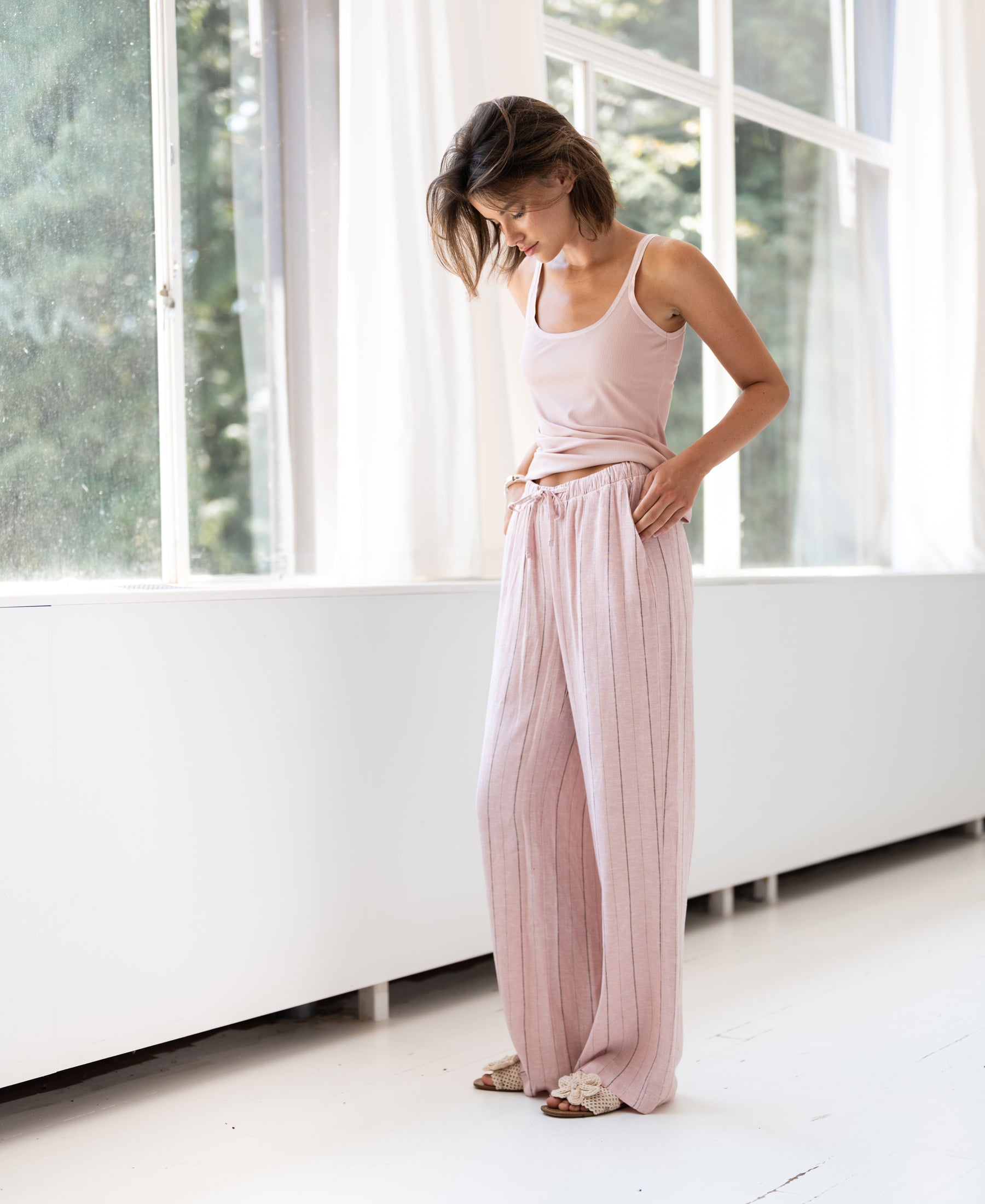 Light pink striped linen-blend pants, model standing by a sunlit window.