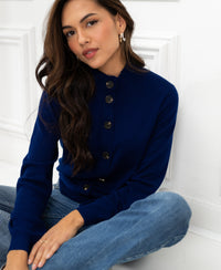 Woman wearing a PLEIN PUBLIQUE Merino wool turtleneck cardigan in navy blue, seated against a white wall.