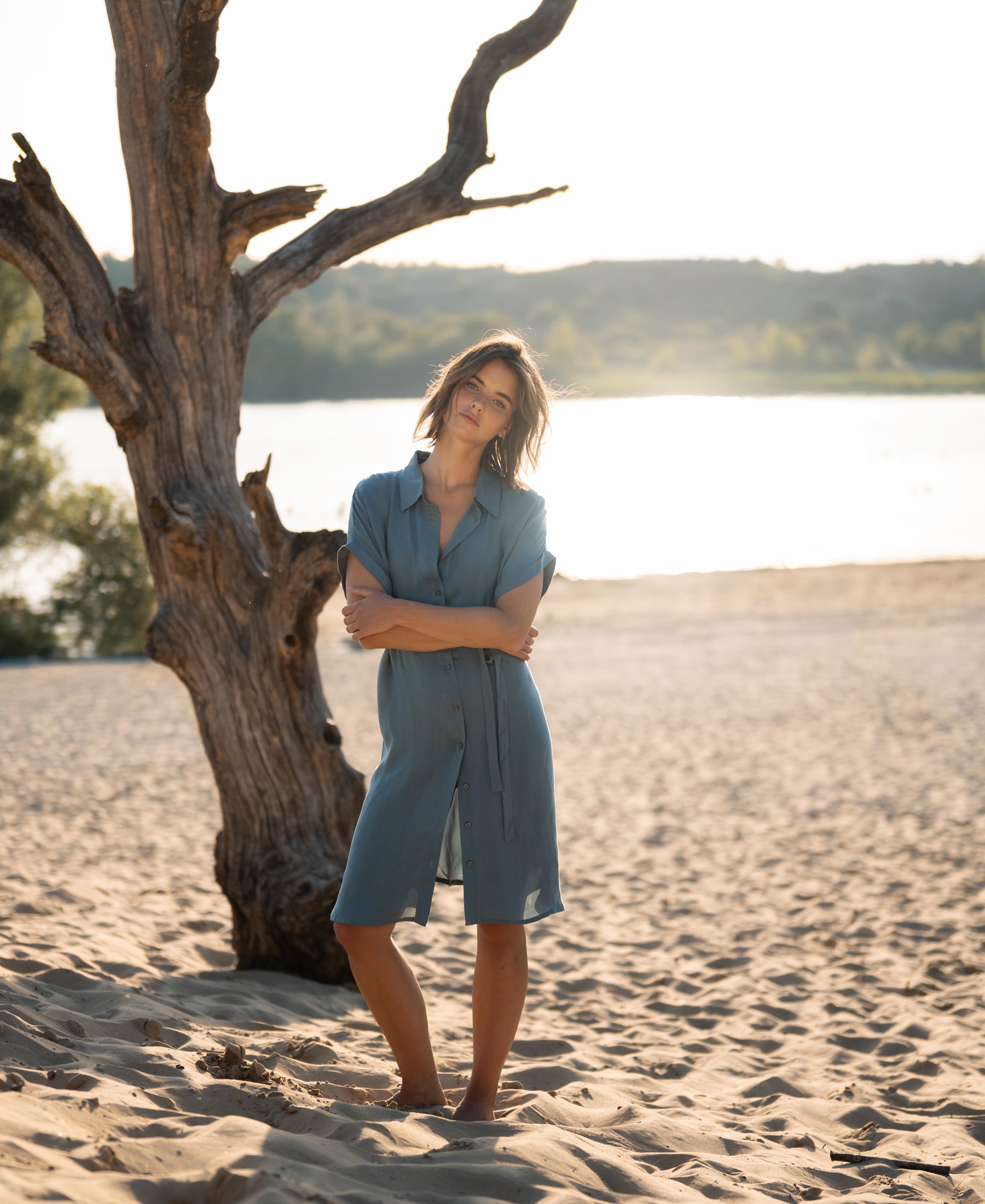 Cupro dress in Denim Cloud blue, shown outdoors with sand, water, and greenery in the background.