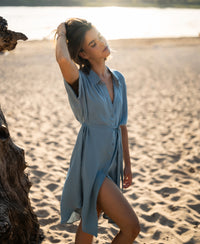 Woman wearing a blue cupro dress on a sandy beach, eyes closed, touching her hair.
