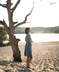 Cupro dress in light blue denim shade, shown on a beach with sunlight glimmering on the water.