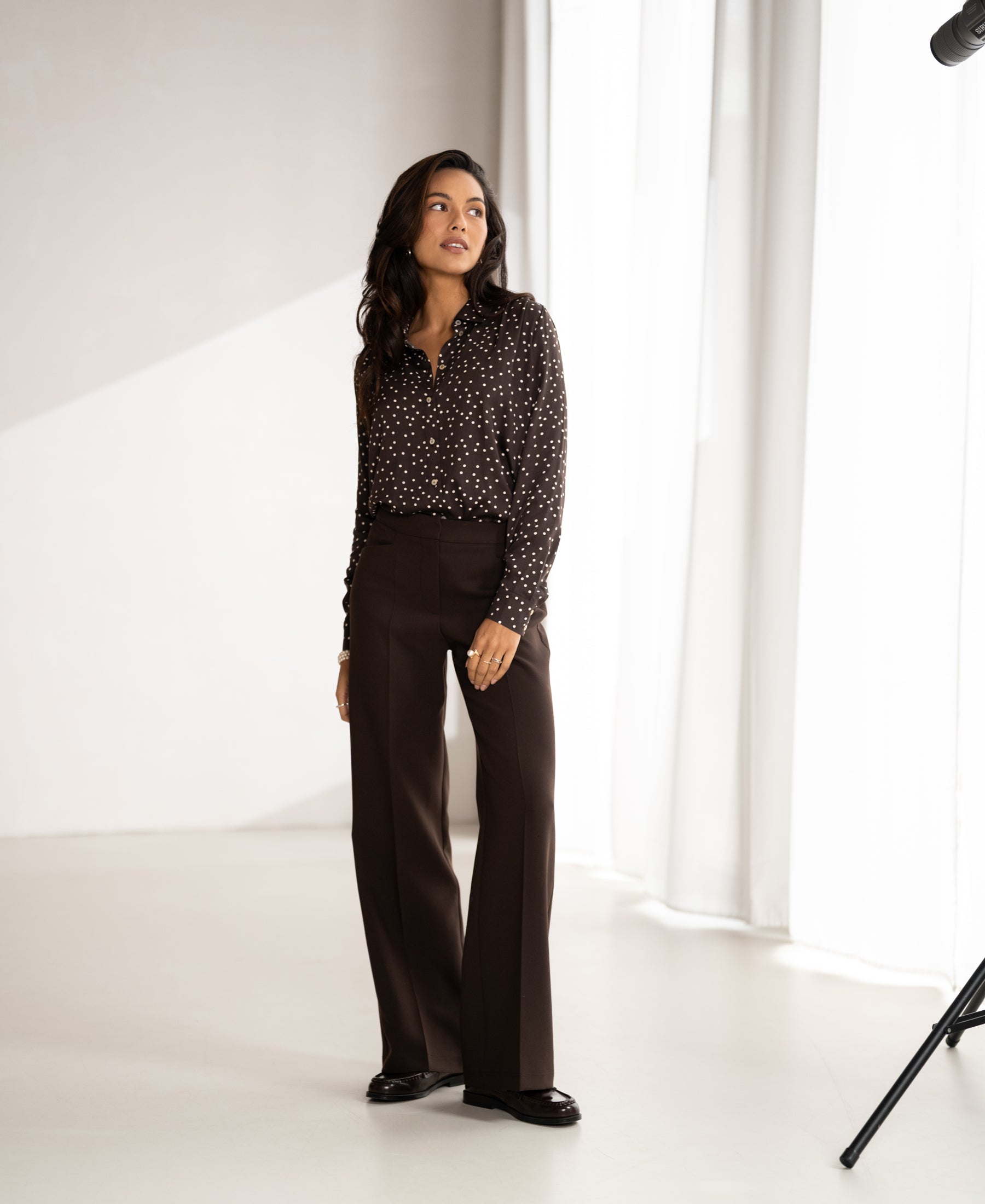 Woman wears a chocolate and ivory polka dot blouse in a bright room with white curtains.