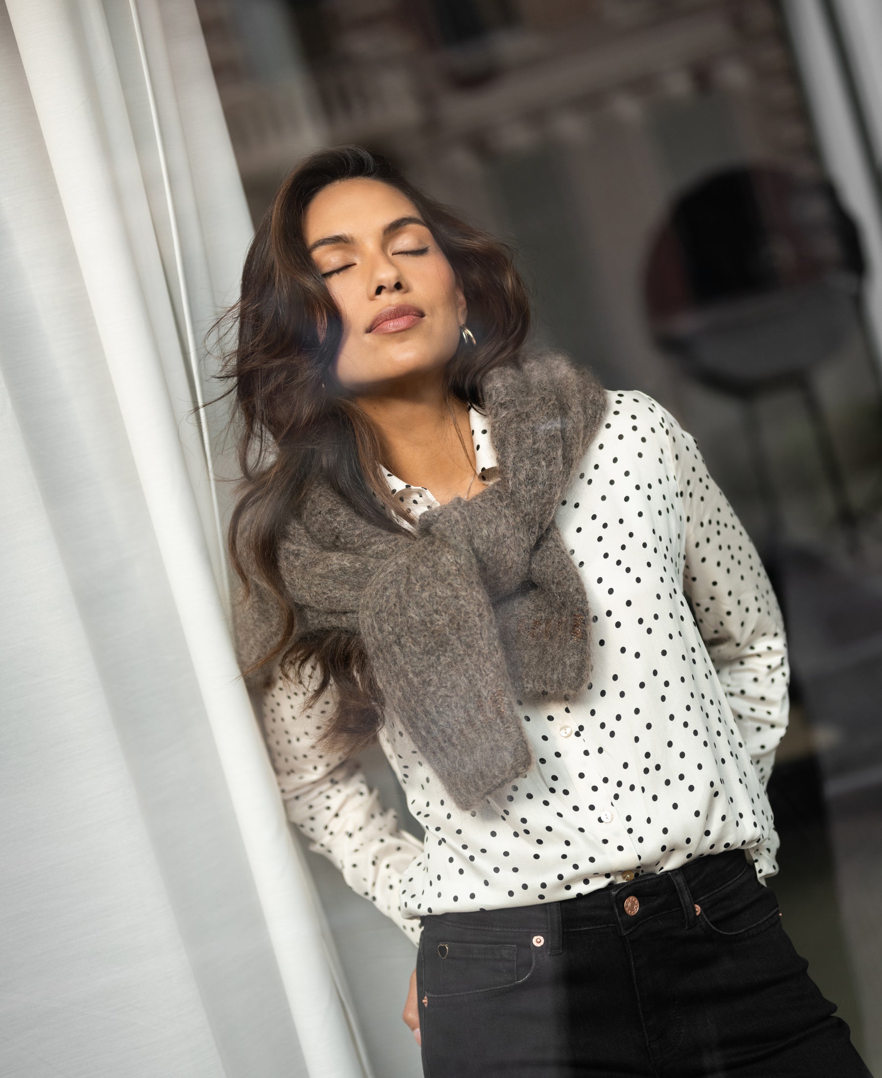 Ivory-black polka dot women's blouse with long sleeves, shown worn by a model standing by a window.