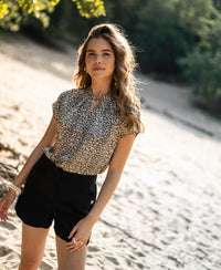 Woman wearing a Plein Publique leopard print blouse top stands outdoors on a sandy path with trees behind her.