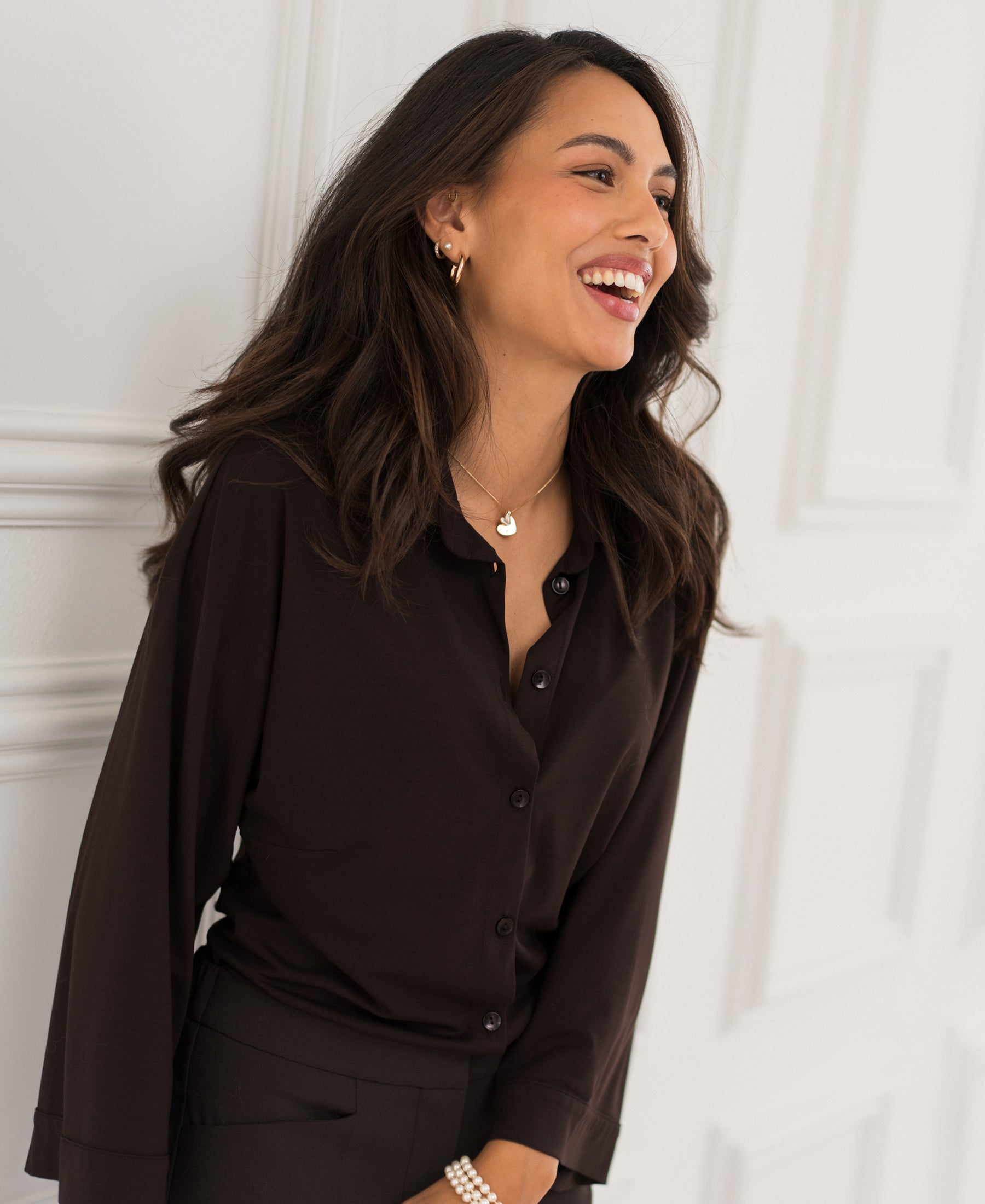 Woman wearing a Plein Publique travel blouse in pure chocolate brown stands against a white wall.