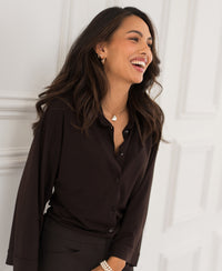 Woman wearing a Plein Publique travel blouse in pure chocolate brown stands against a white wall.