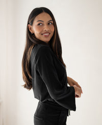 Woman wearing a black PLEIN PUBLIQUE travel blouse, smiling over her shoulder.