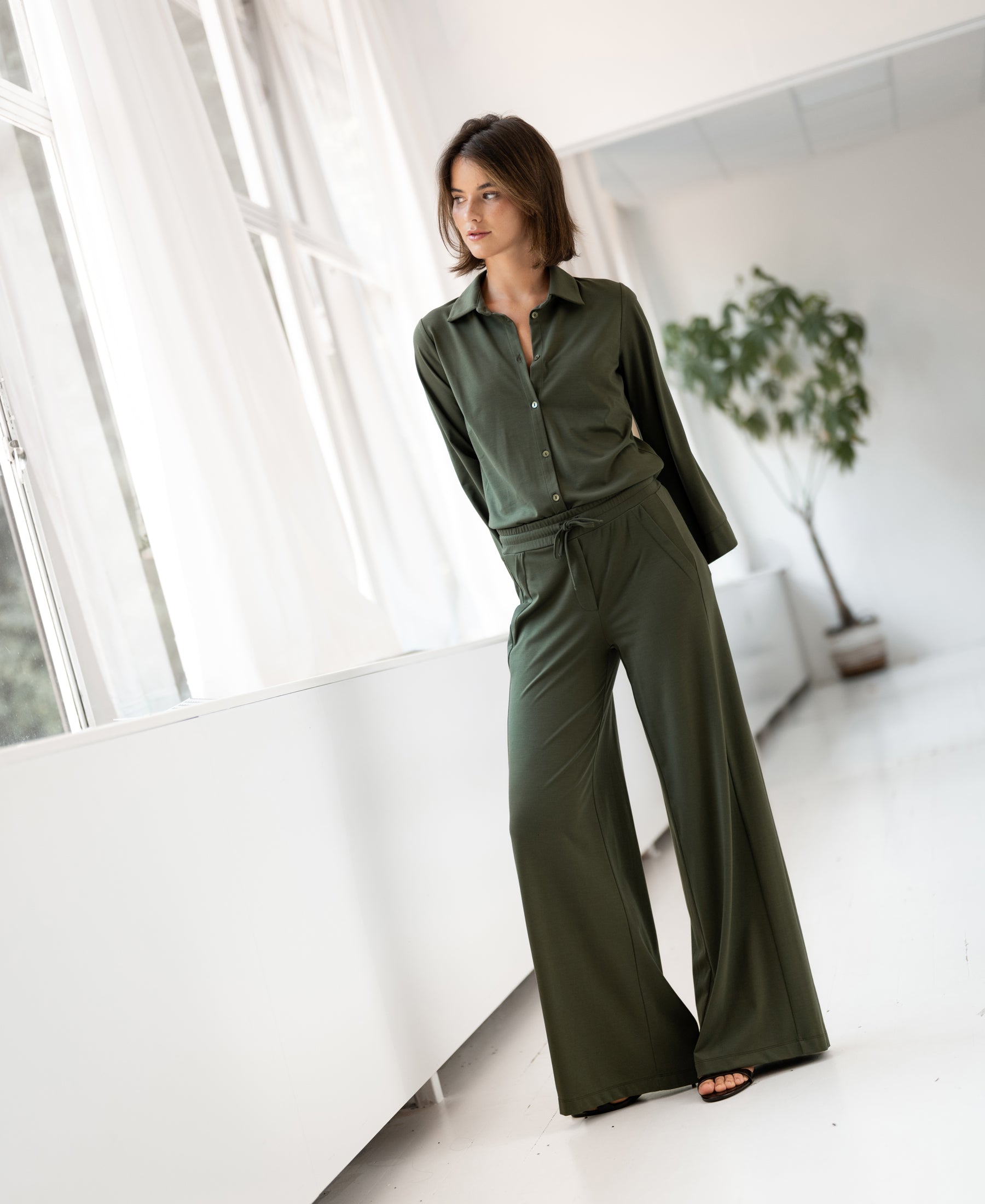 PLEIN PUBLIQUE olive green travel blouse shown indoors near bright windows and a potted plant.