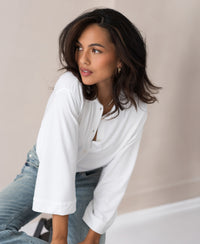 Woman with long dark hair wears a white PLEIN PUBLIQUE travel blouse, looking to the side.