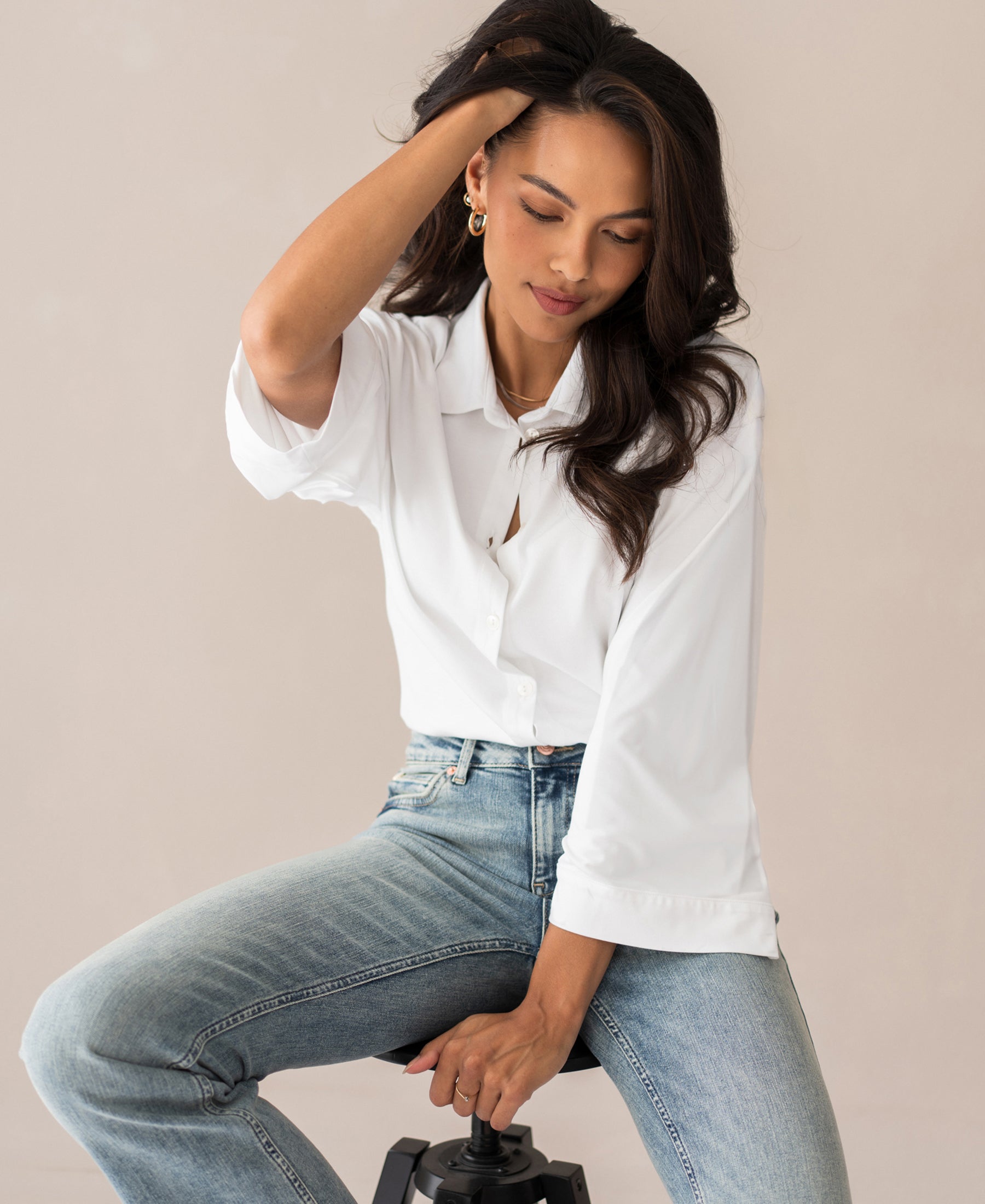Woman wearing white Travelblouse LA SILVESE by PLEIN PUBLIQUE sits on a stool, touching her hair and looking down.