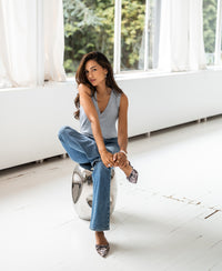Model wearing a gray melange Lyocell top by Plein Publique, seated on a stool in front of large bright windows.