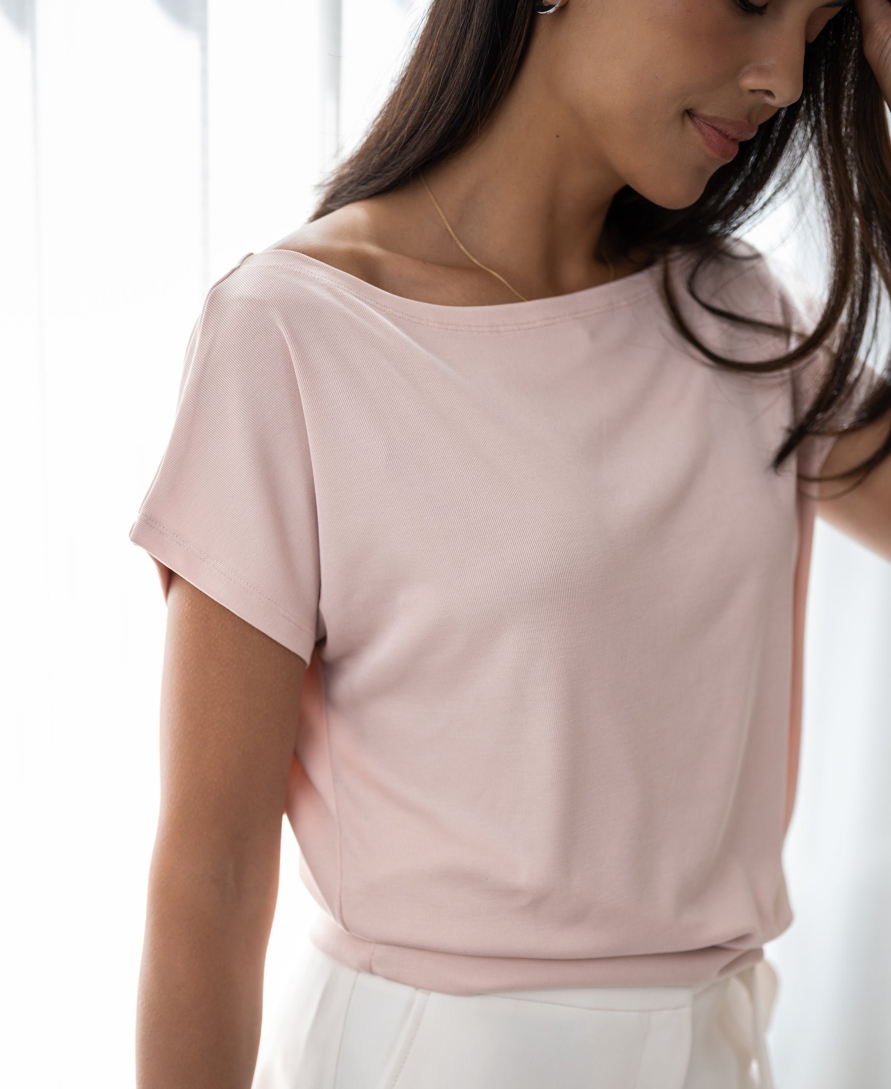 Woman wears the PLEIN PUBLIQUE rib boat neck T-shirt in blush by a sunlit window.