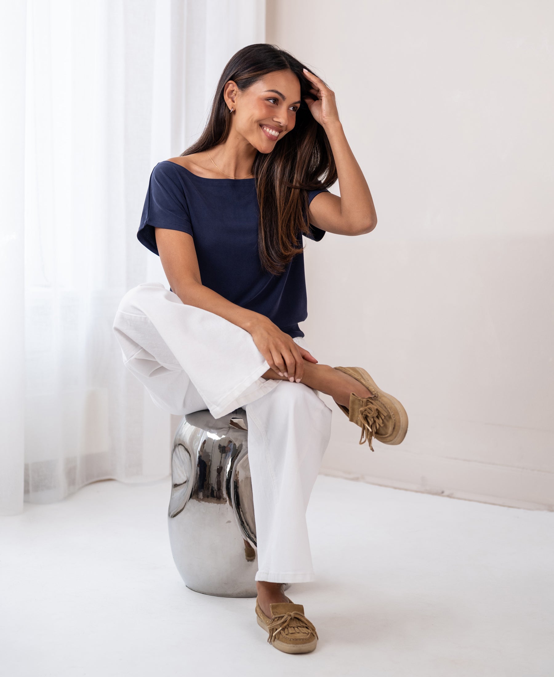 Woman wearing a dark blue ribbed boat neck T-shirt sits on a shiny stool.