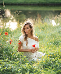 Woman wearing ivory rib boatneck T-shirt sits in tall grass by a pond, holding a red poppy.