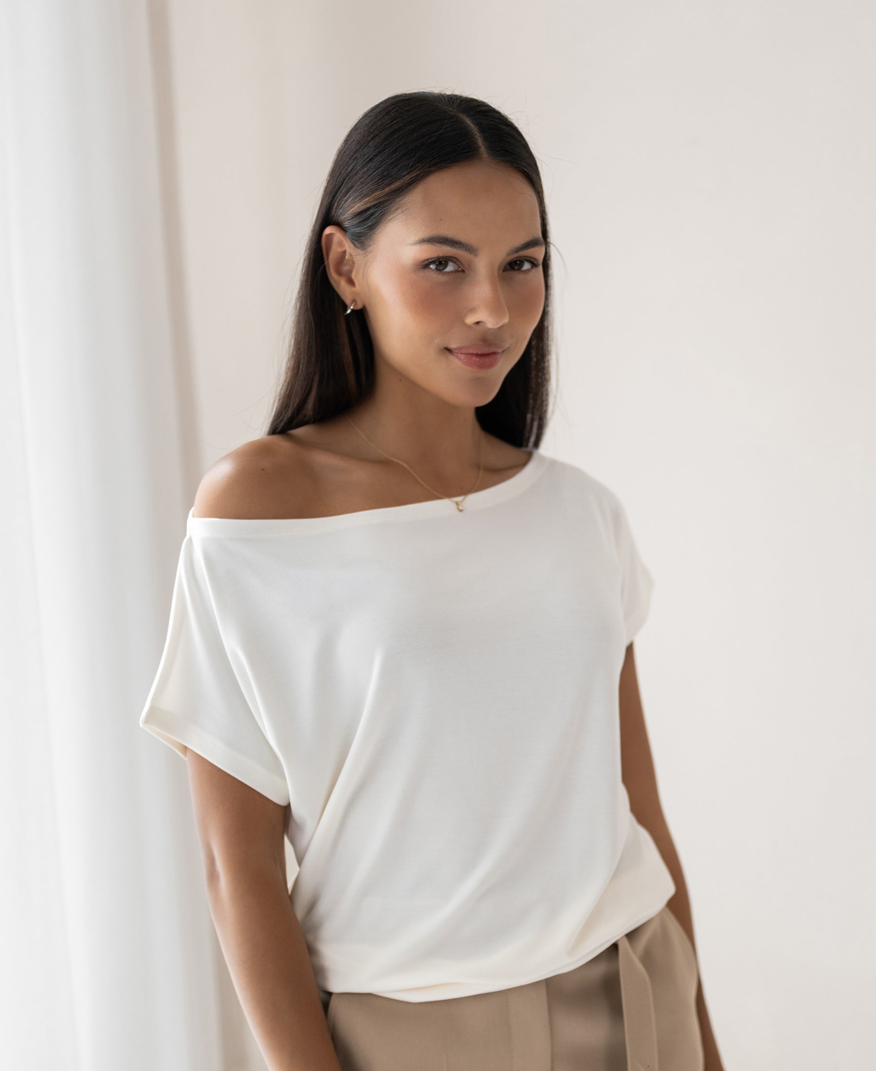 Woman with long dark hair wears an ivory PLEIN PUBLIQUE rib boatneck T-shirt LA SOMME in soft light.