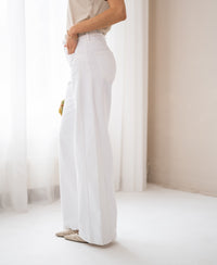 Woman wearing wide leg white jeans stands indoors by a white curtain.