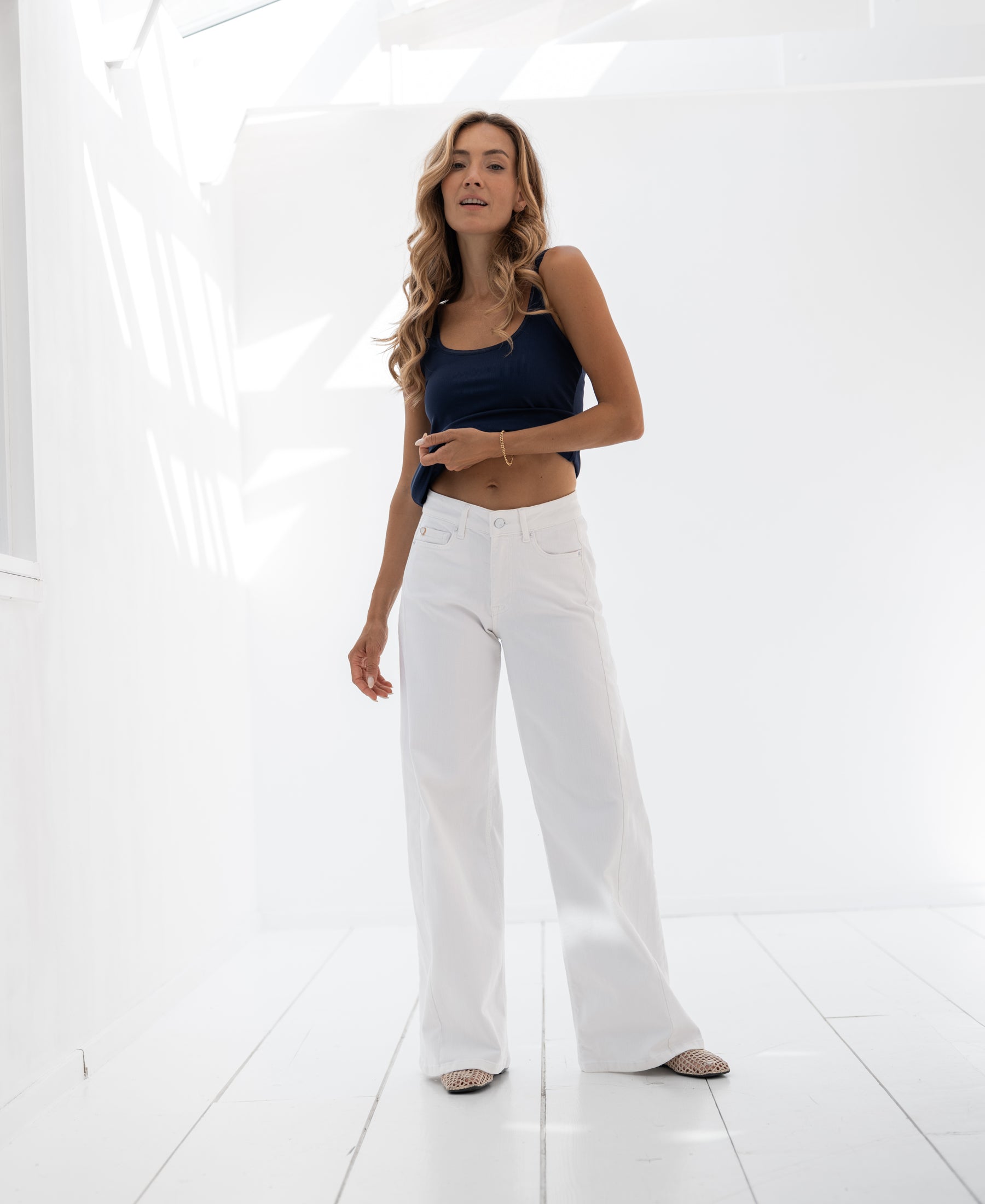Woman wearing white wide leg jeans stands in a bright, sunlit white room.