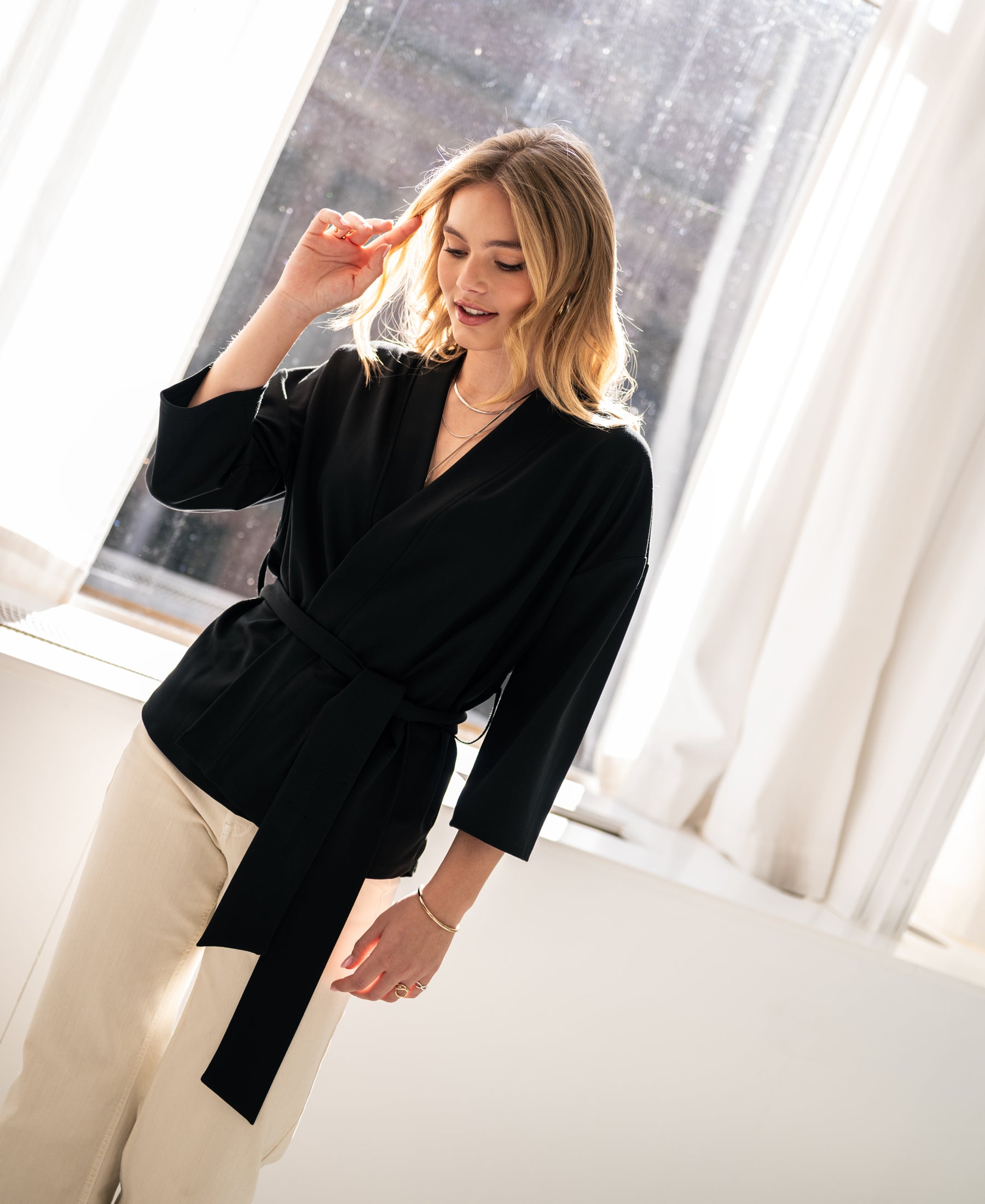 Woman wearing black LA TOKYO Kimono Wrap Jacket by OVERIG stands by a sunlit window, smiling gently.