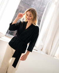 Woman wearing black LA TOKYO Kimono Wrap Jacket by OVERIG stands by a sunlit window, smiling gently.