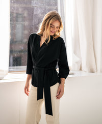 Woman wearing the black LA TOKYO kimono wrap cardigan stands by a sunlit window, smiling.