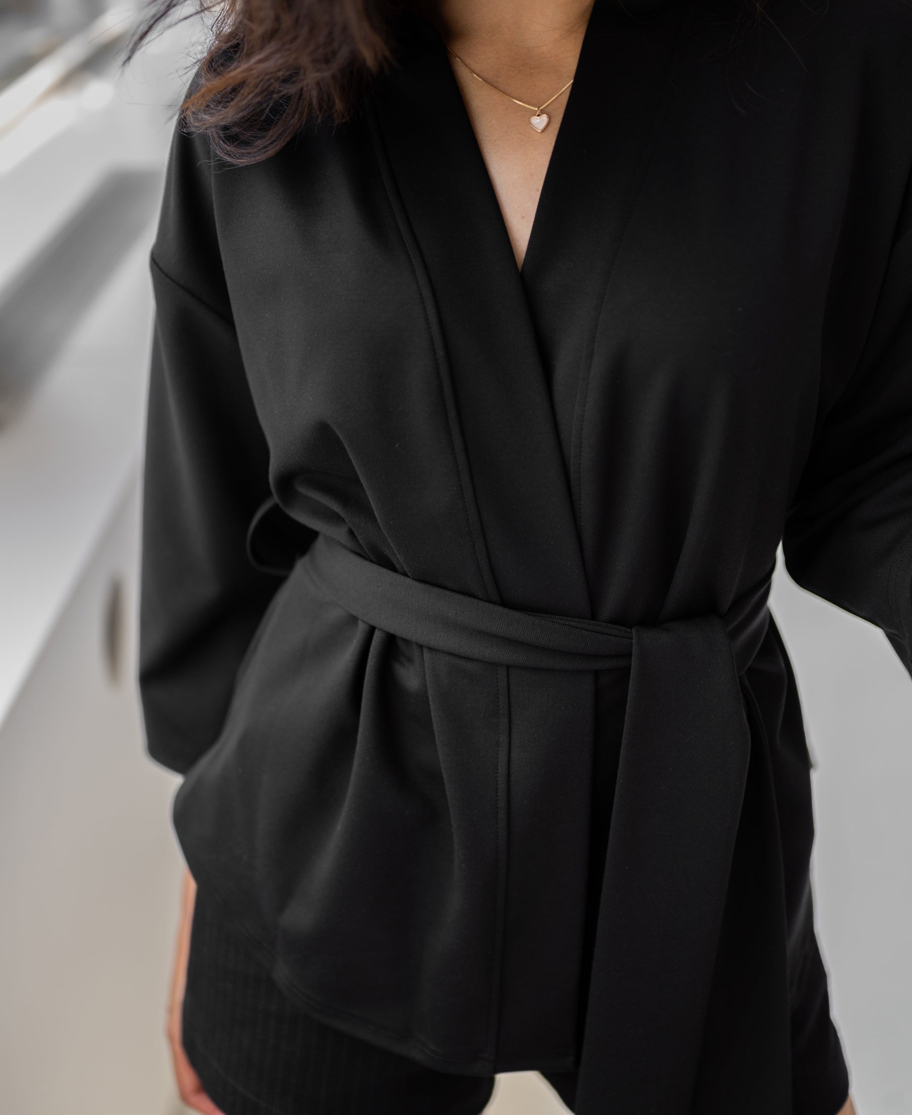Woman wearing a black kimono wrap jacket, standing indoors near a window.