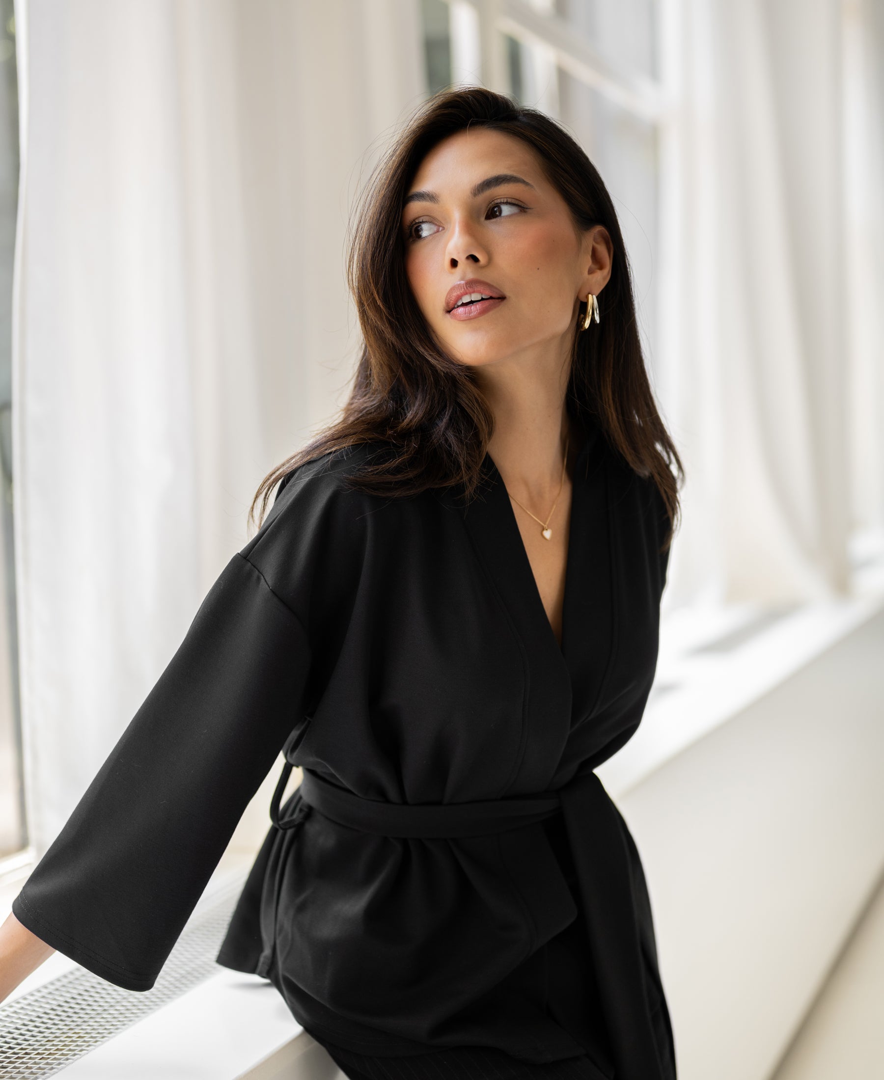 Black LA TOKYO kimono cardigan displayed on a woman sitting by a window with white curtains.