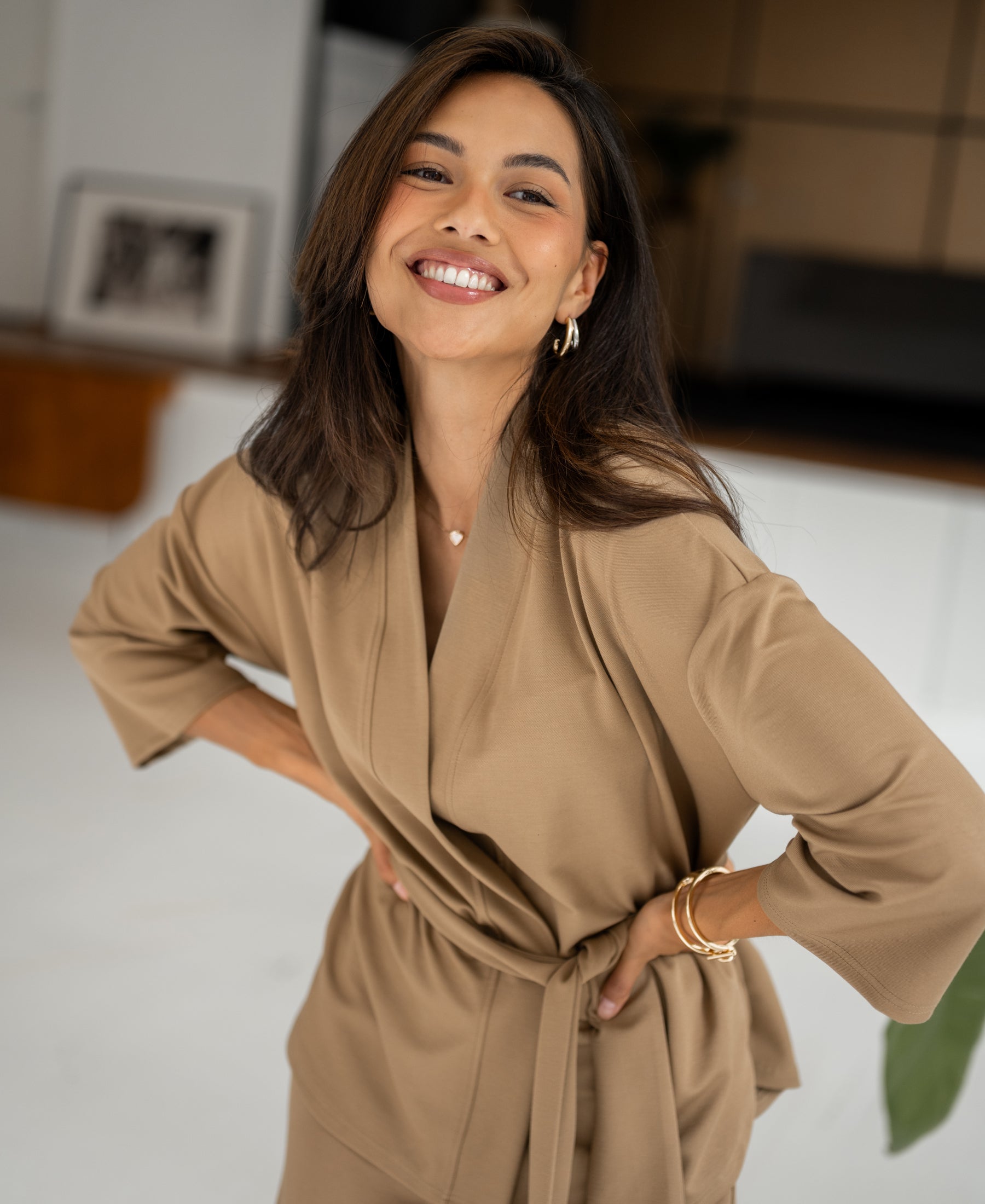 Woman wearing a dark sand kimono wrap jacket stands indoors, hands on hips, blurred background.