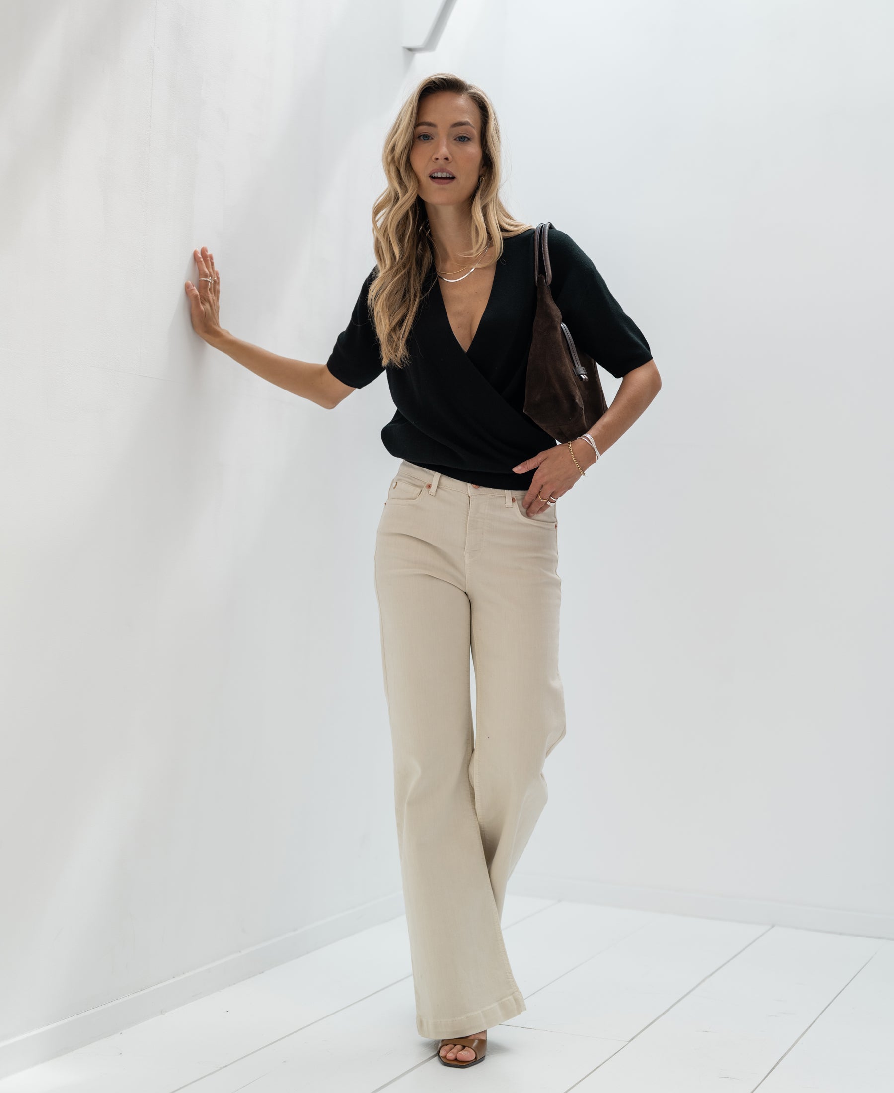 Woman wearing black crossover sweater by PLEIN PUBLIQUE stands against a white wall.