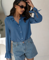 Woman wears a blue PLEIN PUBLIQUE LA TROYES blouse, standing indoors.