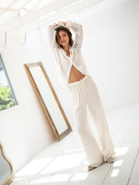Woman wearing cream LA LYNETTE trousers by OVERIG stands in a bright room with a large mirror.