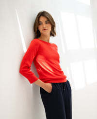 Woman wearing a coral red LA TULIPE boat neck sweater made of Tencel-silk by PLEIN PUBLIQUE, leaning against a white wall.