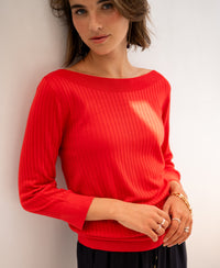 Woman wears coral red Tencel-silk boat neck sweater, standing against a white wall.