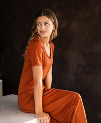 Terra-colored LA FILICIA long dress by Plein Publique shown on a woman sitting on a white surface, side view.